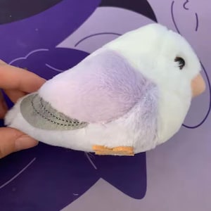 May include: A plush bird toy in white, lavender, and gray. The bird has a small orange beak and black eyes. The wings are gray with stitched detailing. The toy is soft and fluffy, designed to resemble a real bird.