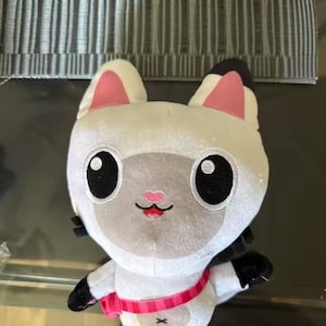 May include: A soft toy cat with a white body, pink inner ears, and black details. The cat has large black eyes, a pink nose, and a pink and white striped collar. The toy is designed to resemble a cartoon character.