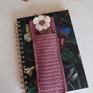 Könnte beinhalten: Ein handgefertigtes, malvenfarbenes Brillenetui aus Häkelarbeit mit einem weißen und malvenfarbenen Blumenakzent. Das Etui ist rechteckig mit einer Tasche für Brillen und kleinen Schlaufen oben. Es liegt auf einem spiralgebundenen Notizbuch mit Schmetterlings- und Libellenmuster.