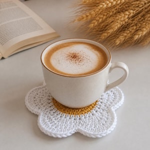 Puede incluir: Una taza de café beige con espuma descansa sobre un posavasos de margarita de ganchillo blanco y amarillo. Un libro y espigas de trigo están en el fondo. La taza tiene un asa blanca.