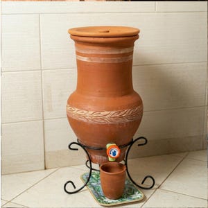 Peut inclure: Un distributeur d'eau en terre cuite avec un couvercle, des bandes décoratives et un robinet. Il repose sur un support en métal noir avec une petite tasse et un plateau. Le distributeur est de couleur marron chaud.