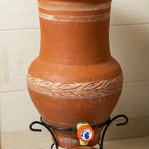 Peut inclure: Un distributeur d'eau en terre cuite avec un couvercle, posé sur un support en métal noir. Le distributeur a un robinet et une tasse assortie. Le corps du distributeur présente des bandes peintes en blanc et un motif de feuilles.