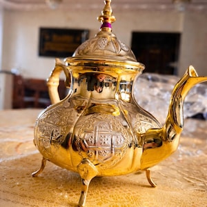 Peut inclure: Une théière dorée avec des motifs en relief complexes. La théière a un bec verseur incurvé, une anse et un couvercle avec un bouton décoratif. Elle repose sur trois petits pieds et est présentée sur une nappe dorée.