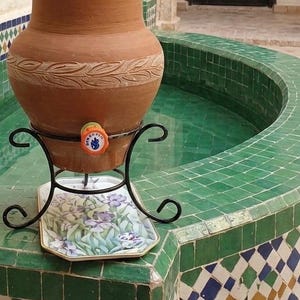 Peut inclure: Un distributeur d'eau en terre cuite avec un motif de feuilles, posé sur un support en métal noir au-dessus d'un plateau floral. Le distributeur a un robinet avec un emblème coloré. Le fond présente une fontaine carrelée verte.