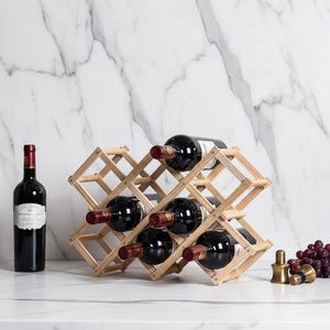 May include: A wooden wine rack holding six bottles of red wine. A full bottle of wine stands to the left. A gold-colored bottle stopper and a bunch of grapes are to the right. The rack is made of light-colored wood.
