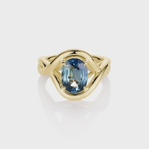 May include: A gold ring featuring an oval-cut, blue-green gemstone. The gemstone is set in a gold band with a unique, interwoven design. The ring is displayed on a white background, highlighting its elegant and modern aesthetic.