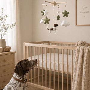 May include: A hanging nursery mobile featuring felt stars in olive green and white, white clouds, and a central brown and white spotted dog figure suspended from a light wood cross frame with small wooden beads on white strings.