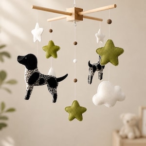 May include: A hanging mobile featuring a wooden cross frame with suspended felt elements including two spotted dogs, three olive green stars, two white stars, a white cloud, and small brown and white felt balls hanging from thin white strings.