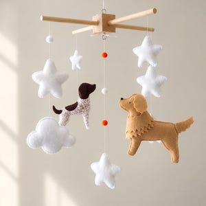 May include: A hanging nursery mobile featuring a light wood cross frame with white felt stars, a white felt cloud, and two felt dog figures. One dog is brown and white with spots, and the other is solid tan. Small orange and white felt balls hang on strings.