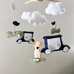 May include: A baby mobile featuring felt cutouts of clouds, cars, and a dog. The mobile has a wooden frame with hanging felt shapes in white, blue, green, and beige. The dog sits on a green cloud with a bone.