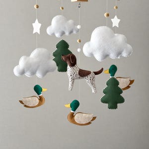 May include: A handmade baby mobile featuring a wooden frame with hanging felt shapes. The mobile includes white clouds, green trees, brown and white spotted dog, ducks, stars, and wooden beads. A charming nursery decoration.