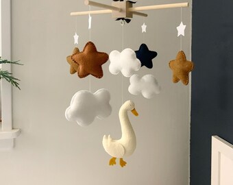 May include: A baby mobile featuring felt stars, clouds, and a goose in shades of brown, white, and navy blue. The mobile is suspended from a wooden frame with a string.