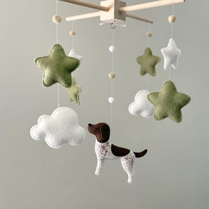 May include: A hanging mobile featuring a central wooden cross frame with white strings suspending felt shapes. The elements include olive green and white stars, white clouds, and a brown and white spotted dog figure, accented with small wooden beads.