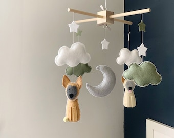 May include: A handmade baby mobile featuring felt clouds, stars, and a crescent moon. The mobile includes two felt dogs in tan and gray, suspended from a wooden frame. The color palette is soft and neutral.