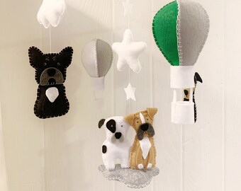 May include: A baby mobile with a gray wooden frame and felt animal and hot air balloon decorations. The mobile features two dogs, a white and black dog and a brown dog, a white cloud, a green and gray hot air balloon, and white stars.