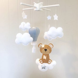 May include: A white wooden baby mobile with a blue hot air balloon, white clouds, a brown teddy bear, and silver stars.