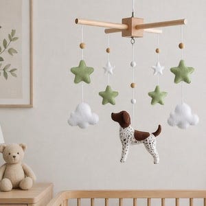 May include: A wooden baby mobile with green stars, white clouds, and a spotted dog. The mobile hangs above a wooden crib with a cream-colored blanket and a cloud-shaped pillow. A framed botanical print and a teddy bear are visible.