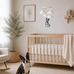 Pu&ograve; includere: Una giostrina per culla con nuvole in feltro bianco e una figura di cane Boston Terrier bianco e nero sospese a una cornice circolare sopra una culla in legno chiaro con una coperta bianca e un cane Boston Terrier seduto sul pavimento.