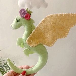 May include: A green felt dragon with pink and purple flowers on its head and yellow felt wings. The dragon is hanging from a white string.