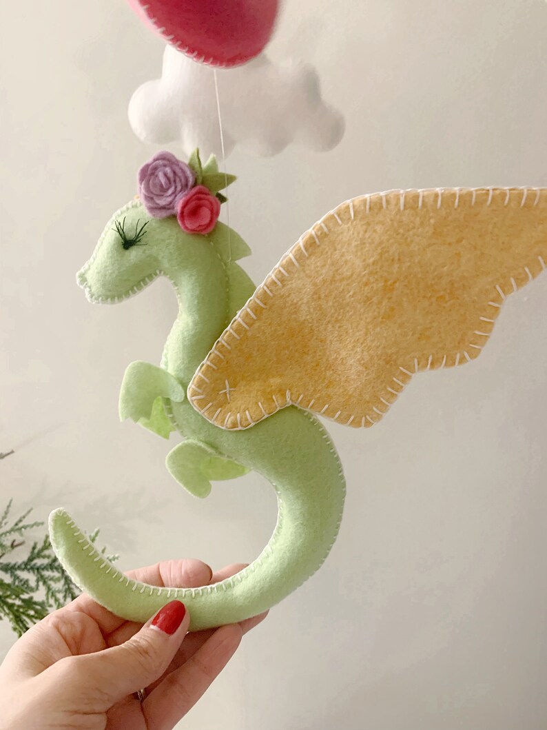 May include: A green felt dragon with pink and purple flowers on its head and yellow felt wings. The dragon is hanging from a white string.