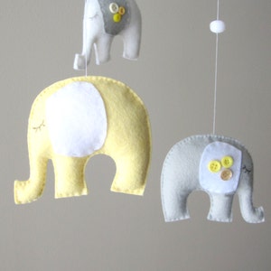 May include: A baby mobile with four felt elephants. Two elephants are yellow with white ears and eyes. Two elephants are grey with white ears and eyes. The grey elephants have yellow buttons on their bodies.