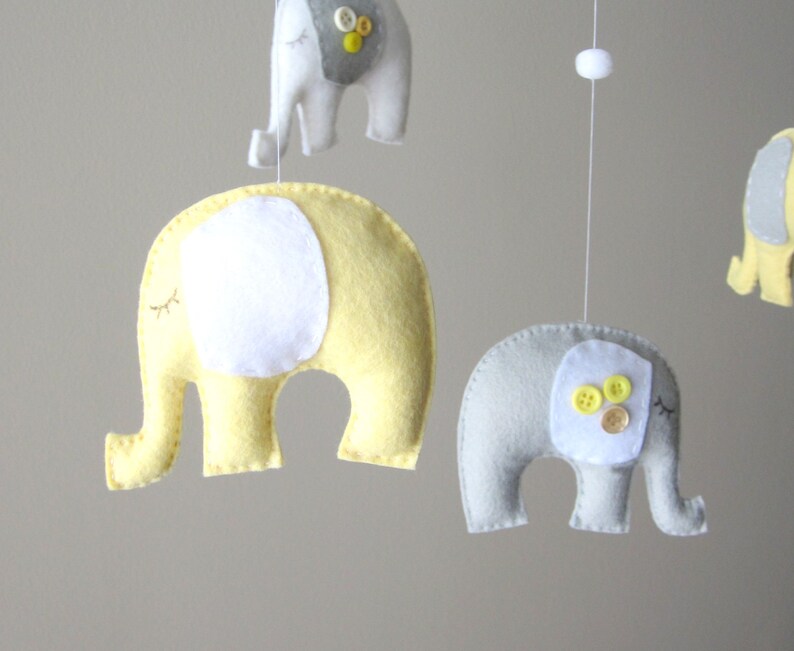 May include: A baby mobile with four felt elephants. Two elephants are yellow with white ears and eyes. Two elephants are grey with white ears and eyes. The grey elephants have yellow buttons on their bodies.