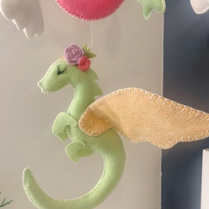 May include: A green felt dragon with pink and purple felt flowers on its head and yellow felt wings. The dragon is hanging from a mobile.