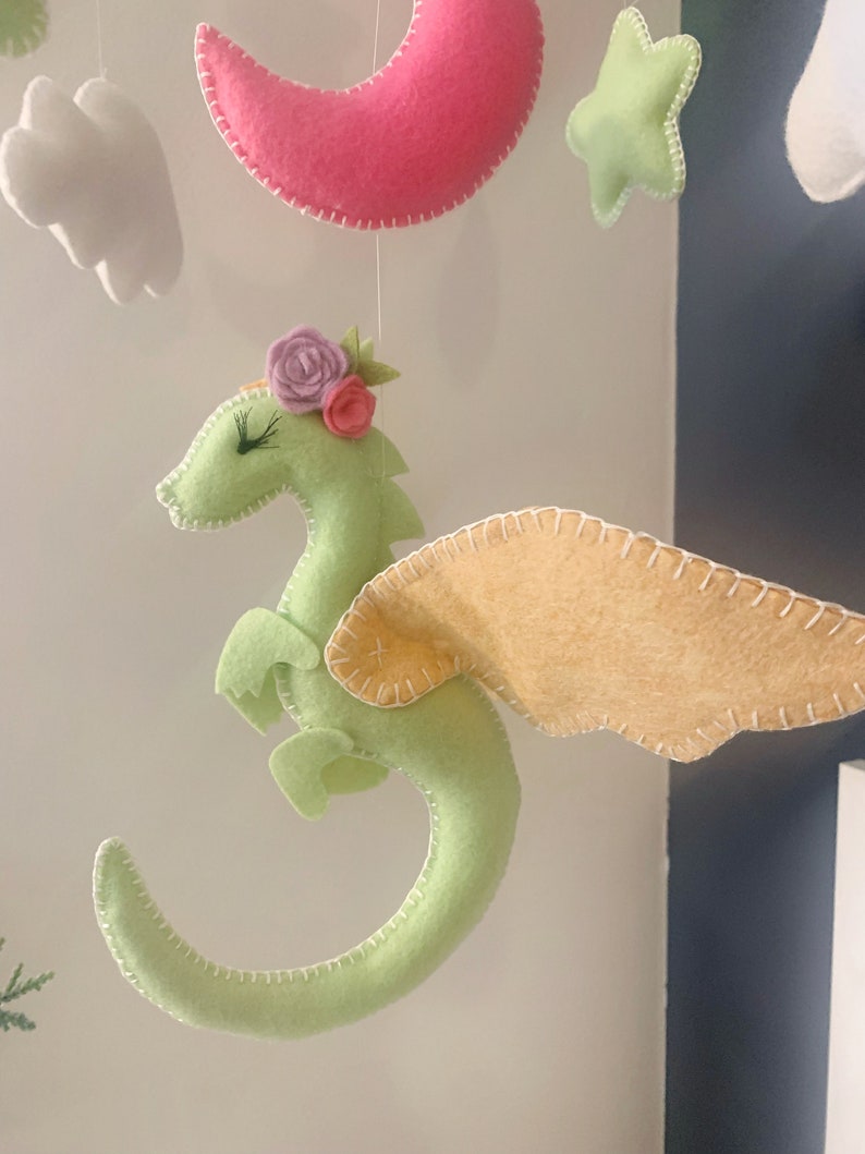 May include: A green felt dragon with pink and purple felt flowers on its head and yellow felt wings. The dragon is hanging from a mobile.