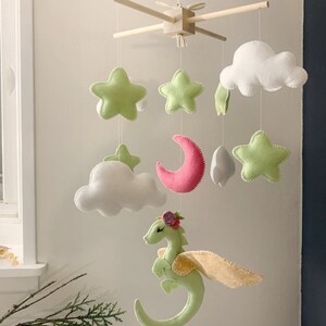 May include: A green felt dragon with pink flowers on its head and yellow wings hangs from a wooden mobile with white clouds, a pink crescent moon, and green felt stars.