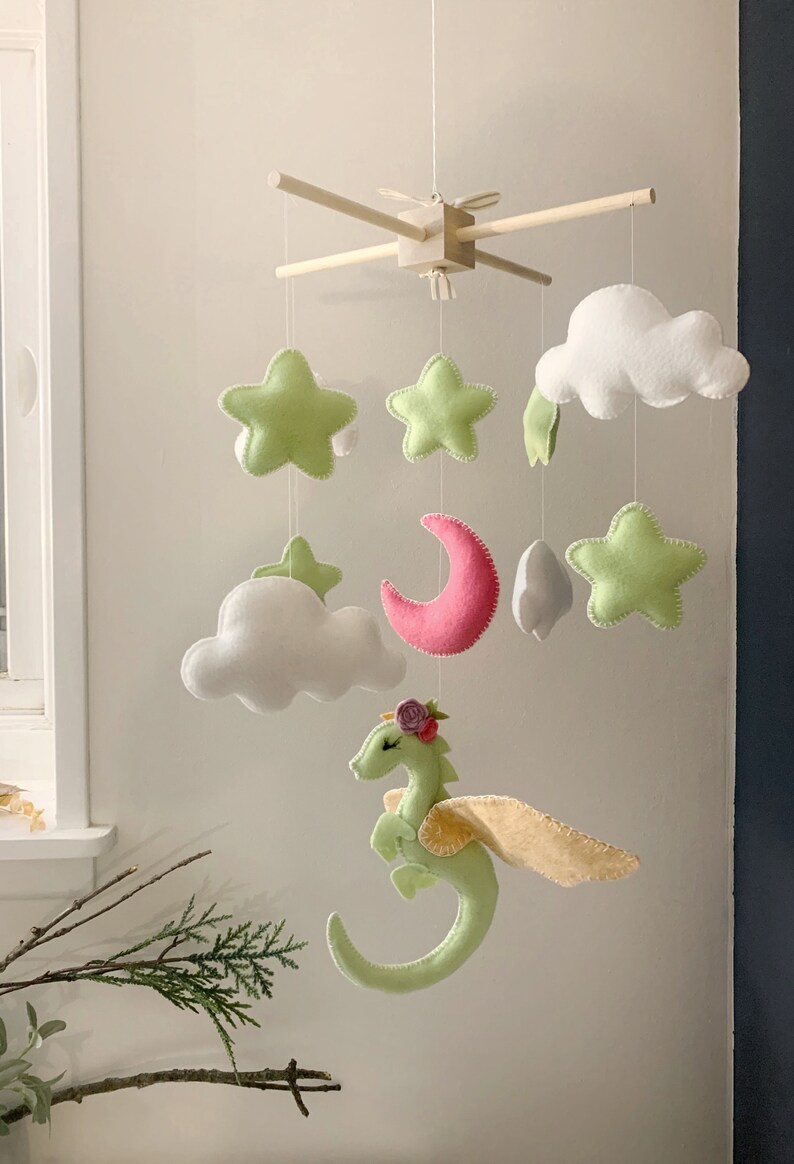 May include: A green felt dragon with pink flowers on its head and yellow wings hangs from a wooden mobile with white clouds, a pink crescent moon, and green felt stars.
