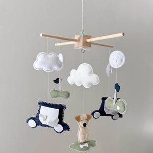 May include: A baby mobile featuring felt cutouts of a dog, cars, clouds, and a moon. The mobile has a wooden frame with hanging felt shapes in white, navy, and green. The dog sits on a green felt patch with a bone.