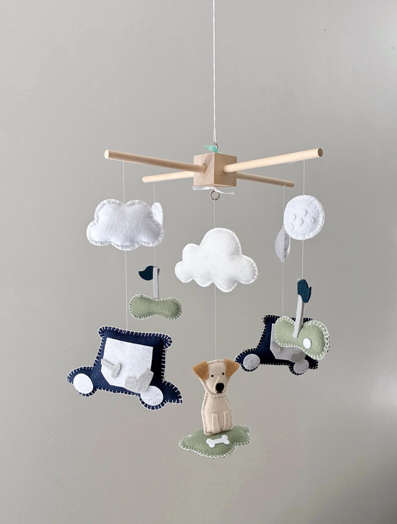 May include: A baby mobile featuring felt cutouts of a dog, cars, clouds, and a moon. The mobile has a wooden frame with hanging felt shapes in white, navy, and green. The dog sits on a green felt patch with a bone.