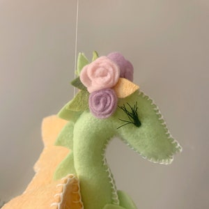May include: A green felt dragon with a yellow felt wing and a pink, purple, and yellow felt flower crown. The dragon has black felt eyelashes.