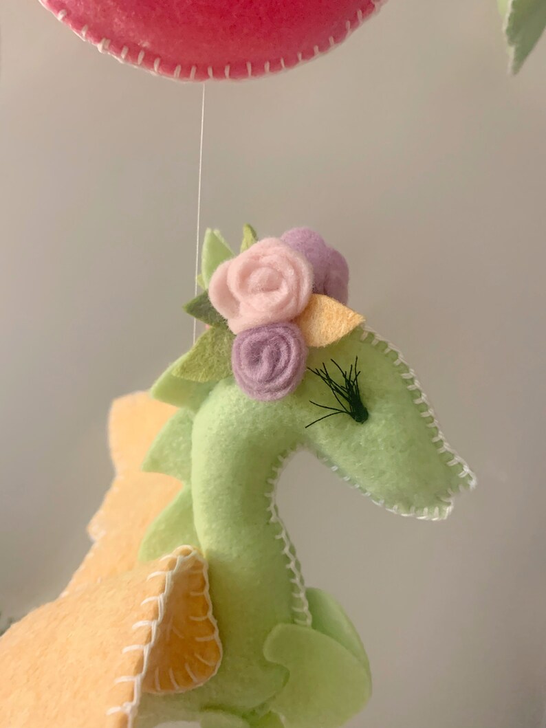May include: A green felt dragon with a yellow felt wing and a pink, purple, and yellow felt flower crown. The dragon has black felt eyelashes.