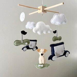 May include: A baby mobile featuring felt cutouts of a puppy, cars, clouds, and a moon. The mobile has a wooden frame with hanging strings. The puppy is beige, and the cars are navy blue and green. The clouds and moon are white.