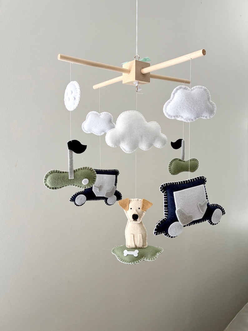 May include: A baby mobile featuring felt cutouts of a puppy, cars, clouds, and a moon. The mobile has a wooden frame with hanging strings. The puppy is beige, and the cars are navy blue and green. The clouds and moon are white.