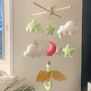 May include: A baby mobile with a green dragon, pink crescent moon, white clouds, and green stars. The mobile is made of felt and is hung from a wooden frame.