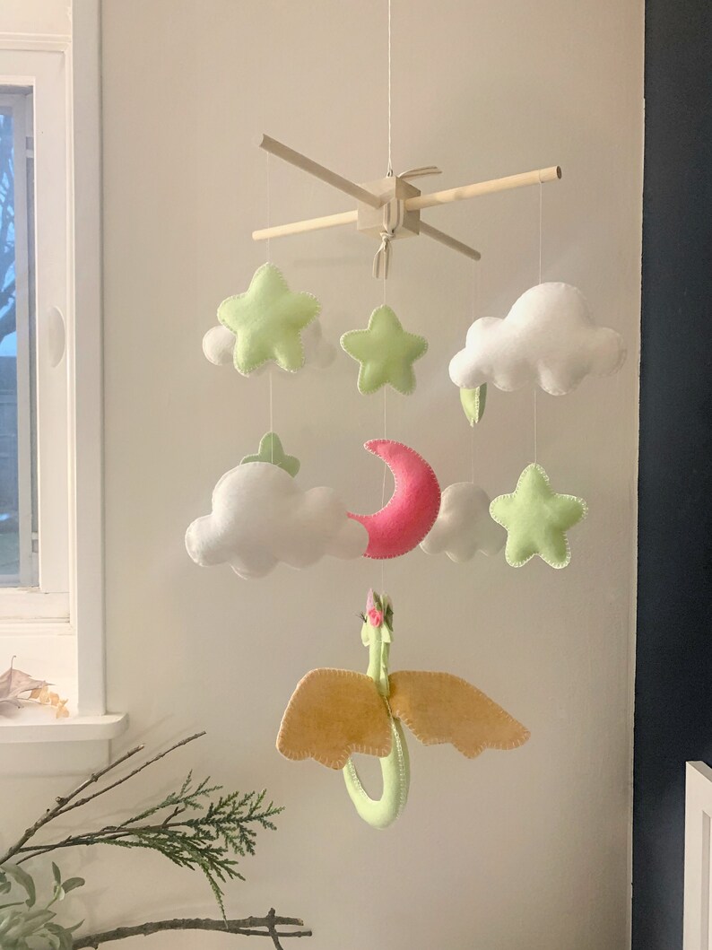 May include: A baby mobile with a green dragon, pink crescent moon, white clouds, and green stars. The mobile is made of felt and is hung from a wooden frame.
