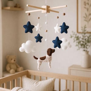 May include: A hanging nursery mobile featuring a light wood cross frame with white strings. It includes felt shapes: navy blue and white stars, white clouds, and a brown and white spotted dog figure, accented with small wooden beads.