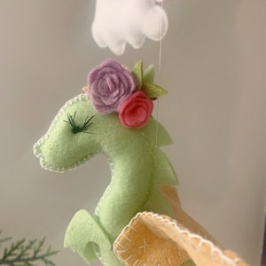 May include: A green felt dragon with pink and purple felt flowers on its head. The dragon has yellow felt wings and white stitching around its edges.