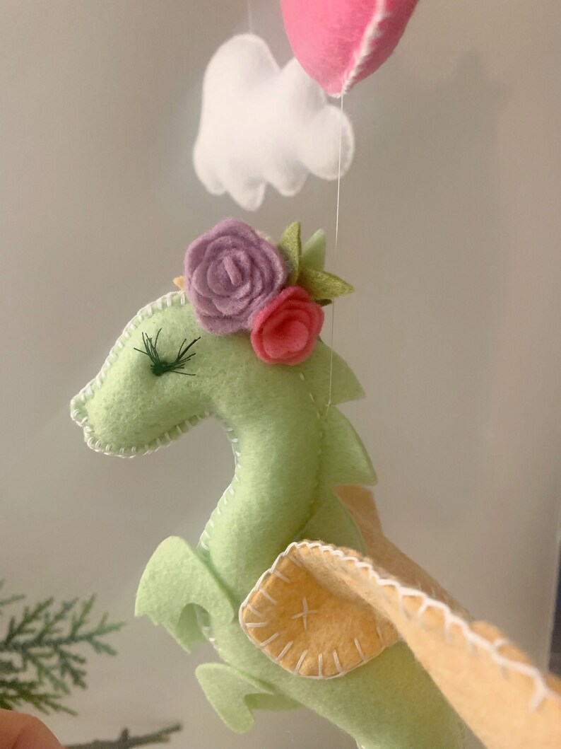 May include: A green felt dragon with pink and purple felt flowers on its head. The dragon has yellow felt wings and white stitching around its edges.