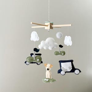 May include: A baby mobile featuring felt cutouts of a dog, cars, clouds, and golf flags. The mobile has a wooden frame with hanging felt shapes in white, green, and navy blue. The dog is beige and the clouds are white.