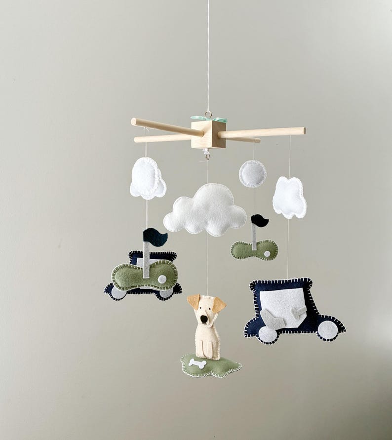 May include: A baby mobile featuring felt cutouts of a dog, cars, clouds, and golf flags. The mobile has a wooden frame with hanging felt shapes in white, green, and navy blue. The dog is beige and the clouds are white.