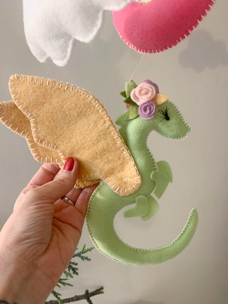 May include: A green felt dragon with yellow felt wings and a flower crown. The dragon is curled up with its tail in its mouth.