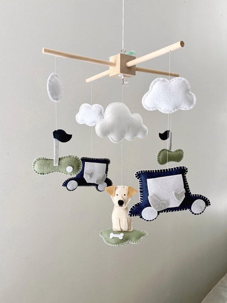 May include: A baby mobile featuring felt cutouts of clouds, cars, and a dog. The mobile has a wooden frame with hanging felt shapes in white, blue, green, and beige. The dog sits on a green cloud with a bone.