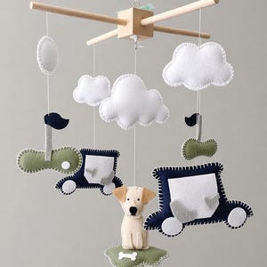 May include: A baby mobile featuring felt cutouts of clouds, cars, and a dog. The mobile has a wooden frame and is suspended by strings. The cars are navy blue and green, and the dog is beige.