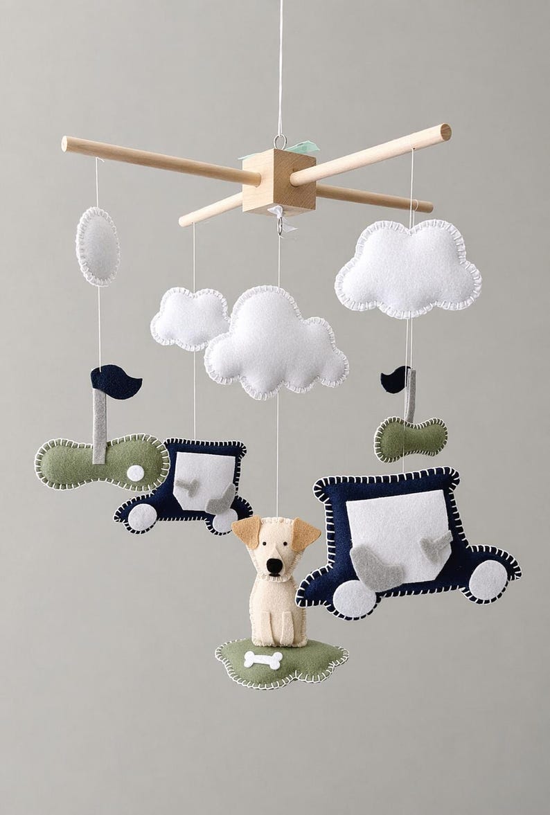 May include: A baby mobile featuring felt cutouts of clouds, cars, and a dog. The mobile has a wooden frame and is suspended by strings. The cars are navy blue and green, and the dog is beige.