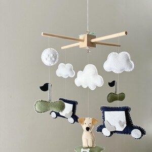 May include: A baby mobile featuring felt cutouts of clouds, a dog, and two cars. The cars are navy blue and green with white accents. The dog is tan and white, and the clouds are white. The mobile is suspended from a wooden frame.