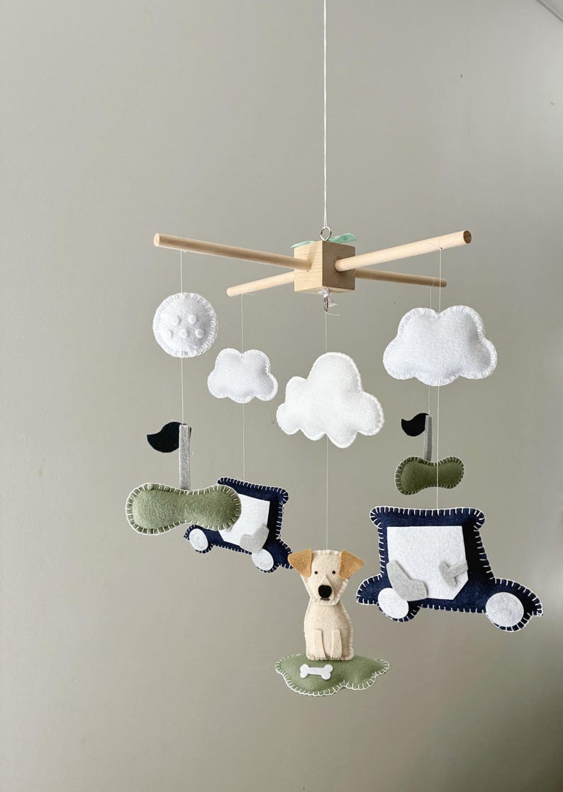 May include: A baby mobile featuring felt cutouts of clouds, a dog, and two cars. The cars are navy blue and green with white accents. The dog is tan and white, and the clouds are white. The mobile is suspended from a wooden frame.