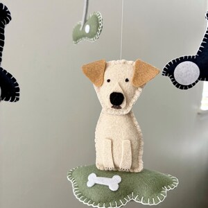 May include: A handmade felt dog mobile featuring a cream-colored dog with tan ears and a black nose. The dog sits on a green cloud with a white bone. Other felt shapes, including a bone and a dark shape, hang above.